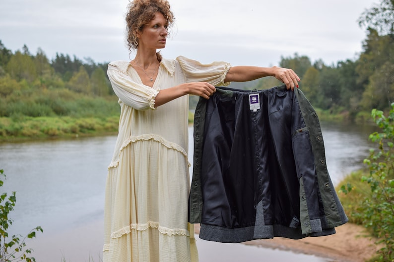 May include: A woman in a white dress holds up a green and black jacket with a purple label. The jacket is unbuttoned and the lining is visible. The woman is standing in front of a river and trees.