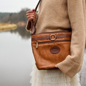 May include: A brown leather crossbody bag with a rectangular shape and a long adjustable strap. The bag features a gold-colored buckle and zipper, and a circular logo. The bag is worn over a tan sweater and white skirt.