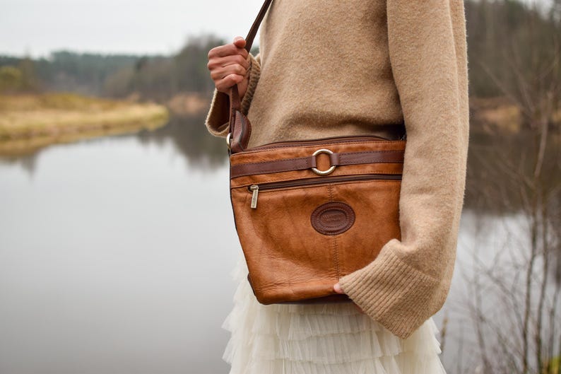 May include: A brown leather crossbody bag with a rectangular shape and a long adjustable strap. The bag features a gold-colored buckle and zipper, and a circular logo. The bag is worn over a tan sweater and white skirt.
