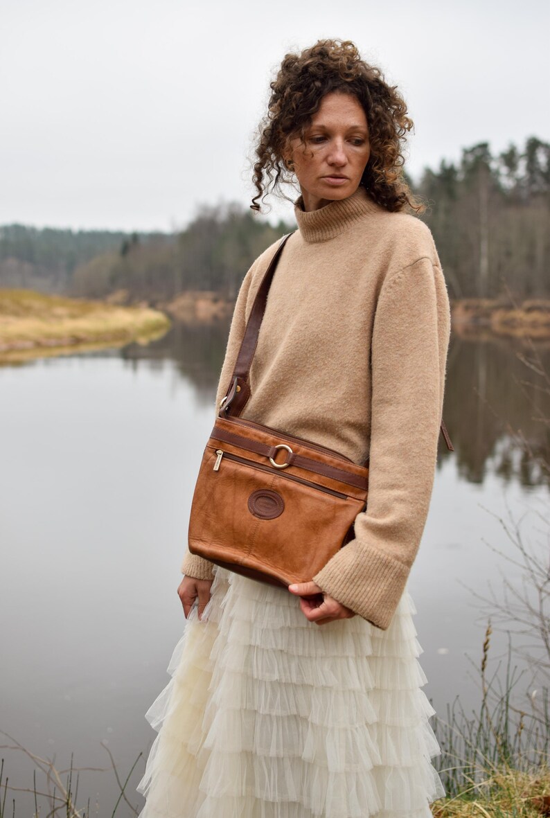 May include: A brown leather crossbody bag with a shoulder strap, a gold-colored ring detail, and a zipper closure. The bag has a circular emblem on the front. The person is wearing a beige sweater and a white skirt.