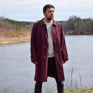 May include: A man wearing a burgundy long-sleeved coat with a brown collar stands by a river. He is wearing a white sweater and dark jeans.