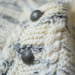 May include: Close-up of a white knitted sweater with two silver buttons. The sweater has a striped pattern with white and gray yarn.