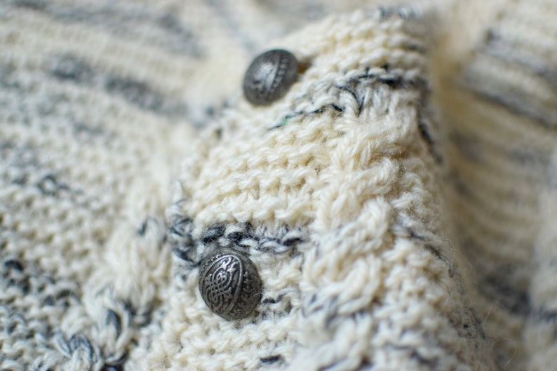 May include: Close-up of a white knitted sweater with two silver buttons. The sweater has a striped pattern with white and gray yarn.