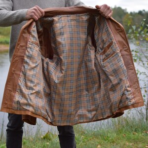 以下が含まれることがあります： 裏地がチェック柄の茶色の革のジャケットが、内側を見せるために開かれています。 ジャケットはボタン留めとポケットが付いたクラシックなデザインです。 裏地は、タン、ブラウン、ブルーのチェック柄です。 ジャケットは屋外で展示されています。