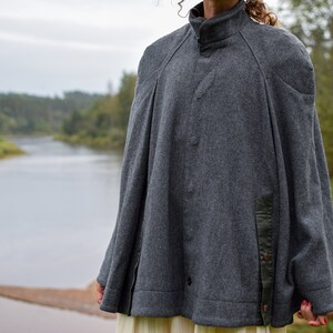 May include: A gray wool cape with a high collar and a button closure. The cape has a black trim on the sides and a black patch on the right shoulder.