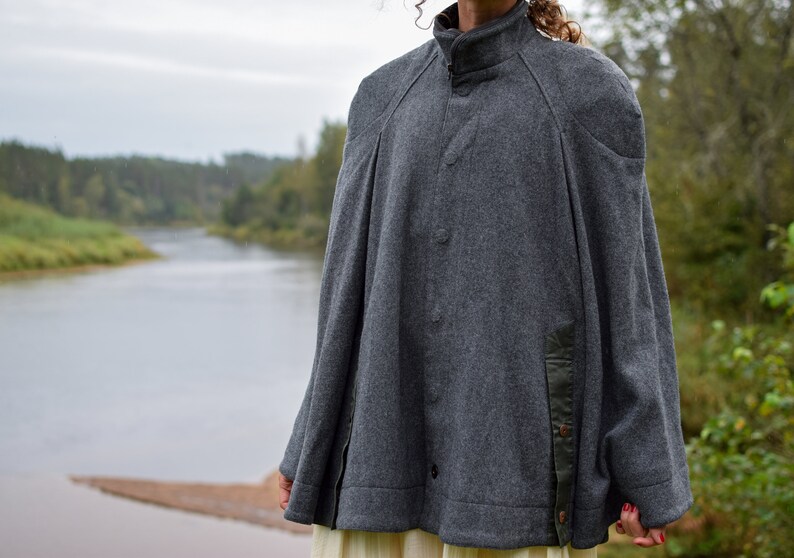 May include: A gray wool cape with a high collar and a button closure. The cape has a black trim on the sides and a black patch on the right shoulder.