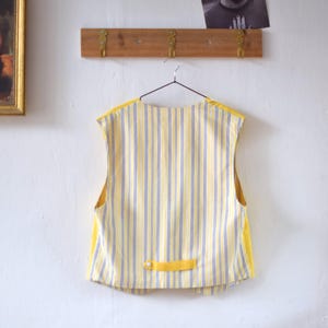 May include: A yellow and blue striped vest with yellow trim hangs on a metal hanger. The vest has a small yellow tab with a button detail at the back. The background includes a wooden rack with brass hooks and a framed picture.