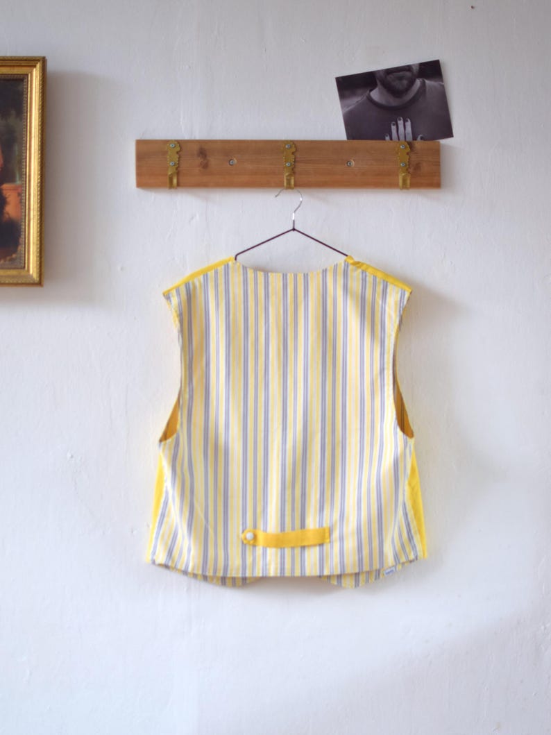 May include: A yellow and blue striped vest with yellow trim hangs on a metal hanger. The vest has a small yellow tab with a button detail at the back. The background includes a wooden rack with brass hooks and a framed picture.