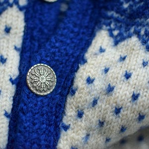 May include: Close-up of a blue and white knit sweater. The sweater features a snowflake button and a pattern of blue diamond shapes on a white background. The knitwear is thick and appears warm.