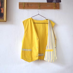 May include: A bright yellow vest with a striped lining and pocket detail. The vest is sleeveless and has a button-up front. The lining features vertical stripes in blue and white. The vest is hanging on a hanger against a white wall.
