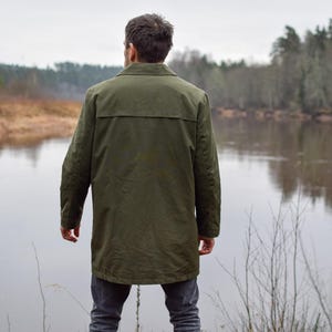 May include: A person wearing a green jacket stands by a river, looking out at the water. The river is surrounded by trees and bushes.