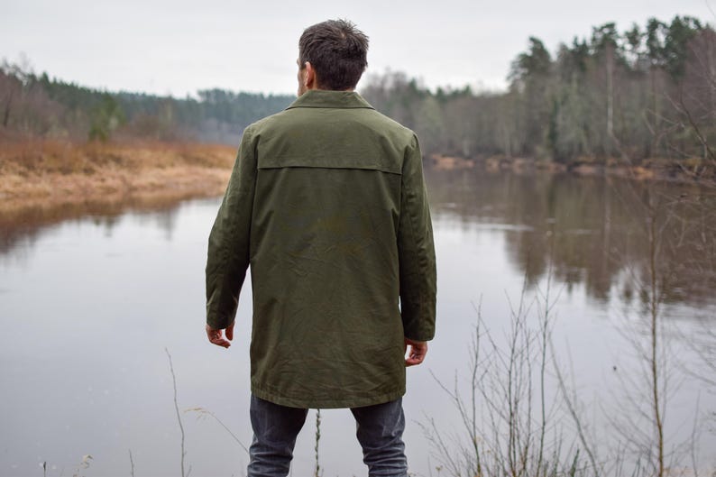 May include: A person wearing a green jacket stands by a river, looking out at the water. The river is surrounded by trees and bushes.