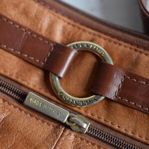 May include: Close-up of a brown leather handbag with a zipper and a gold-colored metal ring. The bag has a stitched design and the zipper pull reads "MADE IN ITALY". The bag is a crossbody or shoulder bag.