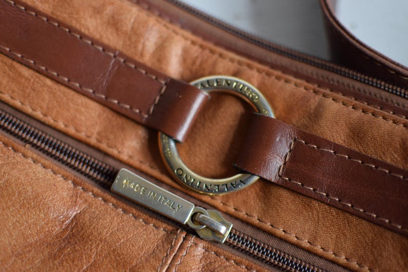 May include: Close-up of a brown leather handbag with a zipper and a gold-colored metal ring. The bag has a stitched design and the zipper pull reads "MADE IN ITALY". The bag is a crossbody or shoulder bag.