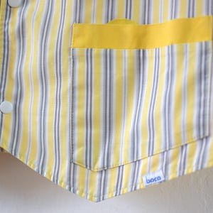 May include: Close-up of a yellow and white striped vest with a yellow pocket. The vest has white buttons and a small blue and white tag that says "boca". The vest is made of a lightweight fabric.