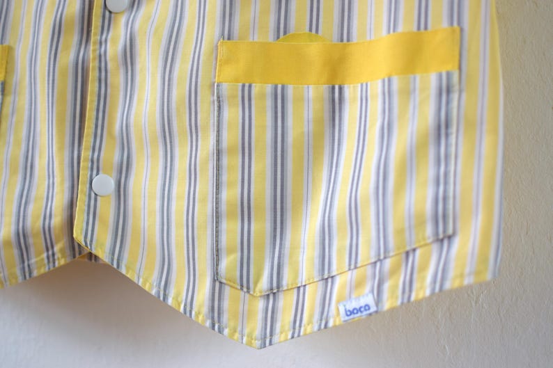 May include: Close-up of a yellow and white striped vest with a yellow pocket. The vest has white buttons and a small blue and white tag that says "boca". The vest is made of a lightweight fabric.