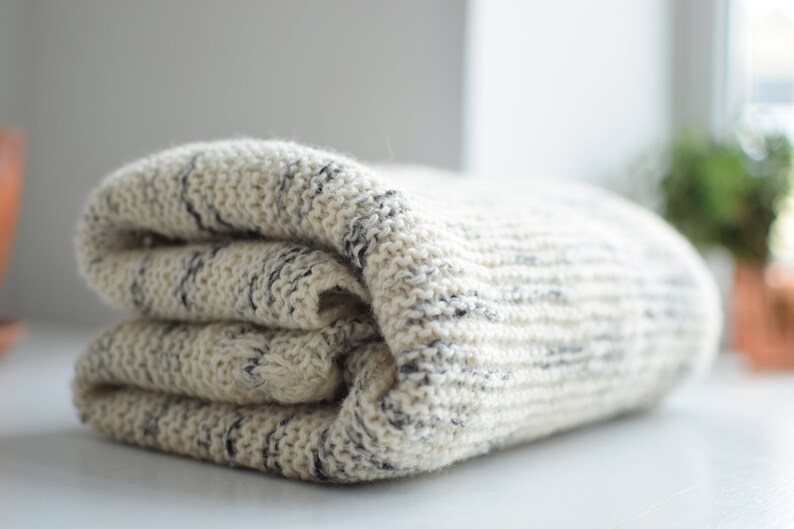 May include: A soft, white and gray knitted throw blanket with a subtle striped pattern. The blanket is folded in thirds and is lying on a white surface.