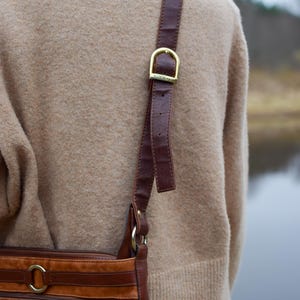 May include: A brown leather crossbody bag with a gold buckle and strap. The bag has a front pocket and a round logo. The strap is attached to the bag with gold rings. The bag is worn over a tan sweater.