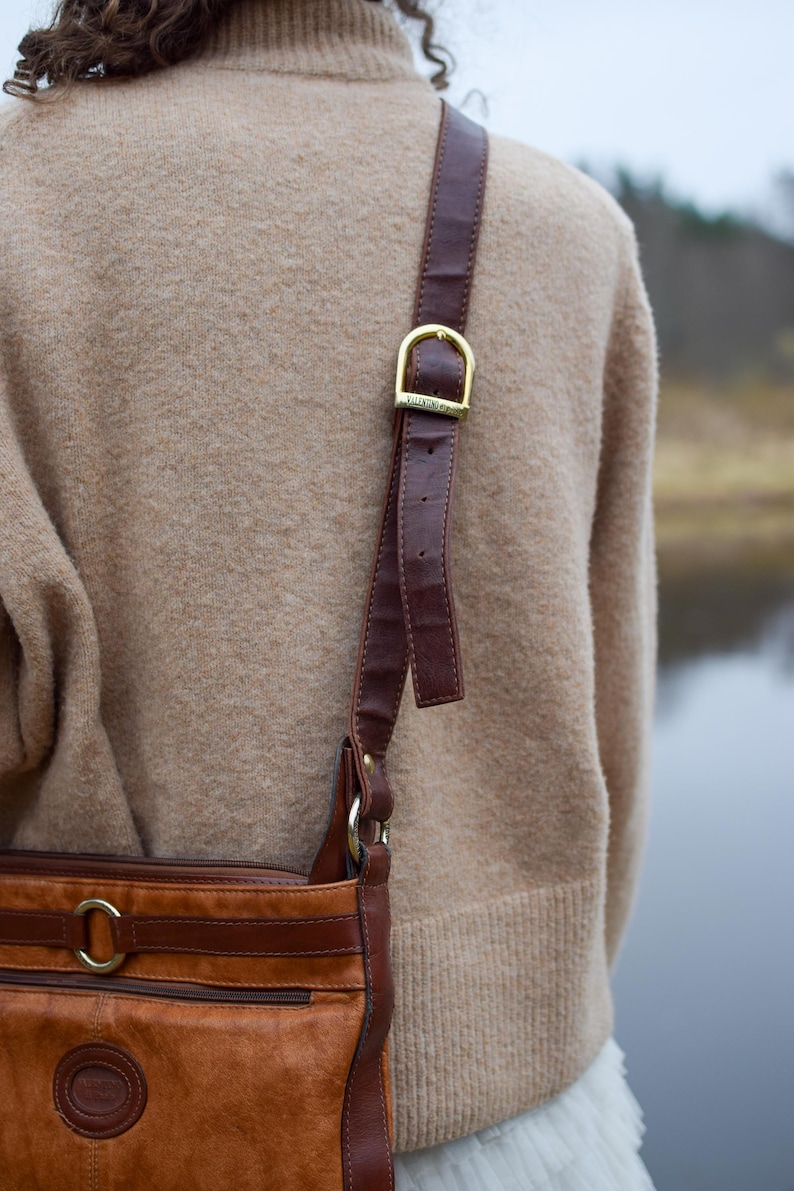 May include: A brown leather crossbody bag with a gold buckle and strap. The bag has a front pocket and a round logo. The strap is attached to the bag with gold rings. The bag is worn over a tan sweater.