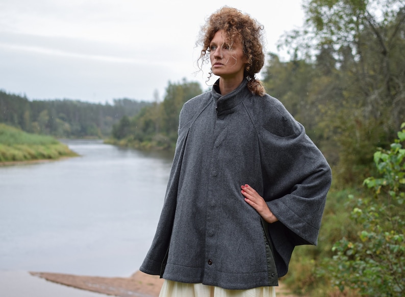 May include: A gray wool cape with a high collar and a button closure. The cape has a relaxed fit and is worn by a person standing in front of a river and trees.
