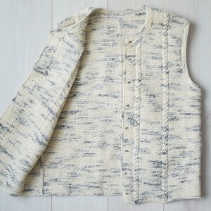 May include: A white knitted vest with a cable knit design and a button closure. The vest has a speckled pattern with blue and white yarn.