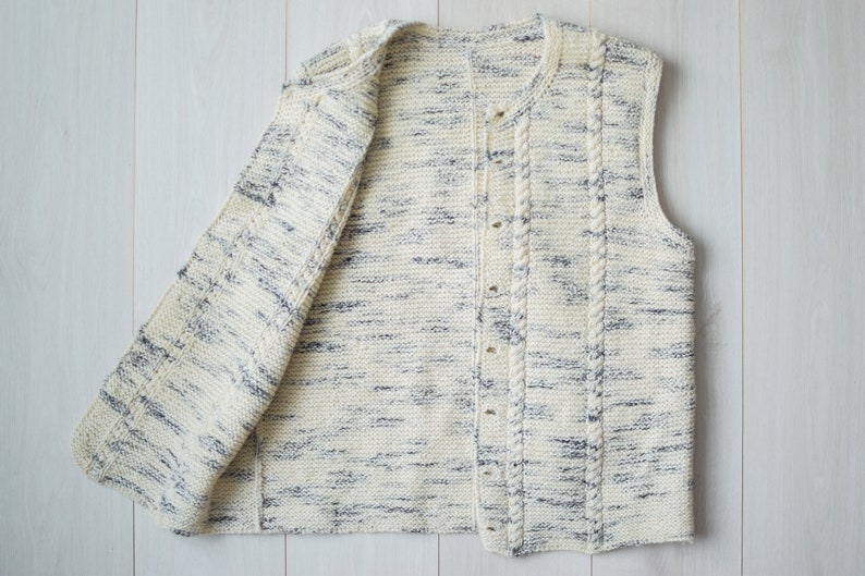 May include: A white knitted vest with a cable knit design and a button closure. The vest has a speckled pattern with blue and white yarn.