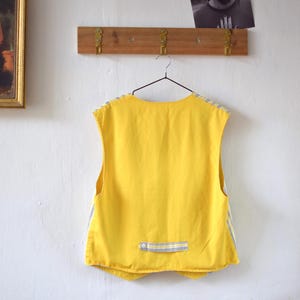 May include: A sleeveless yellow vest with blue and white striped accents hangs from a metal hanger. The vest has a small pocket with a blue and white striped detail. The vest is hanging on a white wall.