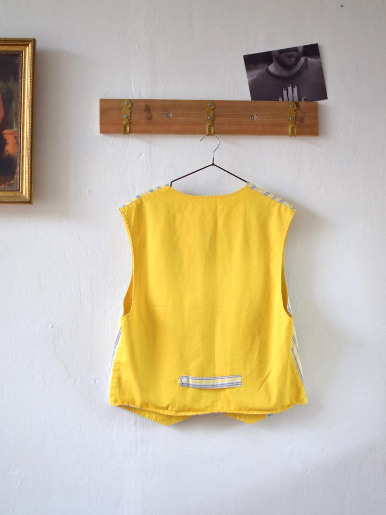 May include: A sleeveless yellow vest with blue and white striped accents hangs from a metal hanger. The vest has a small pocket with a blue and white striped detail. The vest is hanging on a white wall.