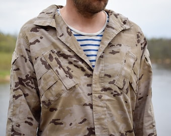 May include: A tan and brown camouflage jacket with multiple pockets. The jacket is worn over a white and blue striped shirt. The background shows a body of water and trees.