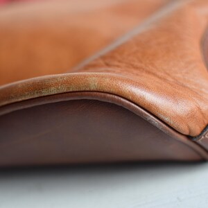 May include: Close-up of a tan leather bag with a dark brown leather base and trim. The bag's leather has a textured appearance, with visible grain and slight color variations. The stitching is visible along the edges.