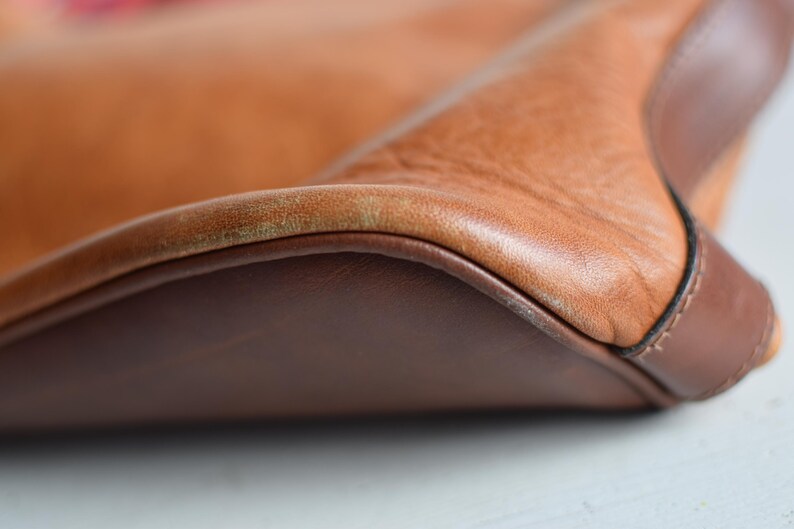 May include: Close-up of a tan leather bag with a dark brown leather base and trim. The bag's leather has a textured appearance, with visible grain and slight color variations. The stitching is visible along the edges.