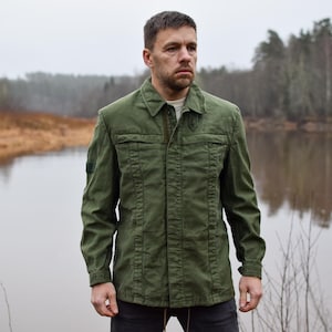 Könnte beinhalten: Olivgrüne Jacke mit Knopfleiste, zwei Brusttaschen und Kragen. Die Jacke wird über einem hellen Hemd getragen. Der Hintergrund zeigt ein Gewässer und Bäume.