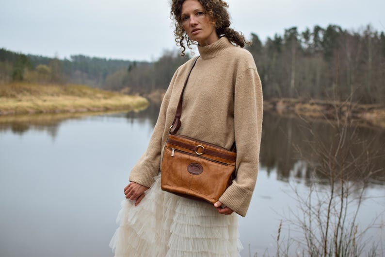 May include: A brown leather crossbody bag with a gold-tone buckle and zipper detail. The bag has a shoulder strap and a logo on the front. The person is wearing a beige sweater and a white skirt.