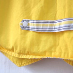May include: Close-up of a bright yellow fabric with a striped accent. The accent features a white button and a gray, white, and yellow striped pattern. The fabric appears to be part of a garment or textile item.