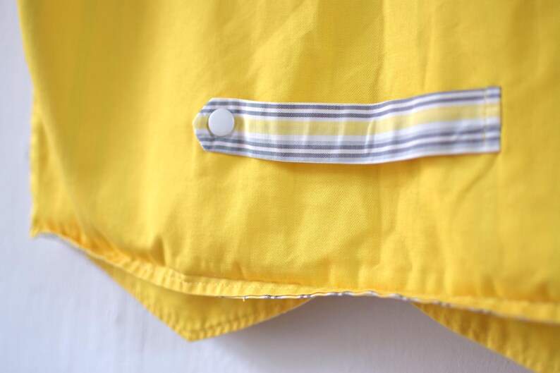 May include: Close-up of a bright yellow fabric with a striped accent. The accent features a white button and a gray, white, and yellow striped pattern. The fabric appears to be part of a garment or textile item.