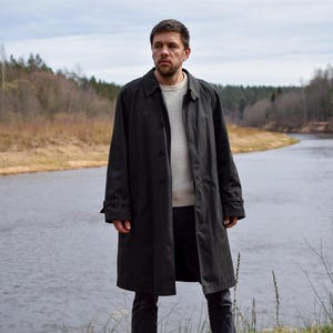 May include: A man wearing a black long-sleeved coat stands in front of a river. He is wearing a white shirt underneath the coat. The river is surrounded by trees and bushes.