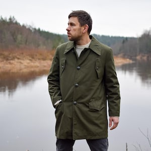 May include: A man wearing a green, long-sleeved, button-up jacket stands by a river. He is looking to the left of the frame. The river is calm and the water is reflecting the trees on the opposite bank.