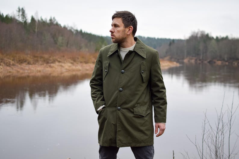 May include: A man wearing a green, long-sleeved, button-up jacket stands by a river. He is looking to the left of the frame. The river is calm and the water is reflecting the trees on the opposite bank.