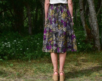 May include: A knee-length skirt with a floral pattern in shades of purple, yellow, and white on a dark base. The skirt is worn with platform sandals. The person is standing in a woodland setting.