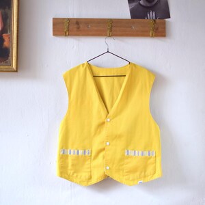 May include: A bright yellow vest with a V-neck and three white buttons. The vest has two front pockets with blue and white striped accents. The vest is hanging on a black hanger against a white wall.
