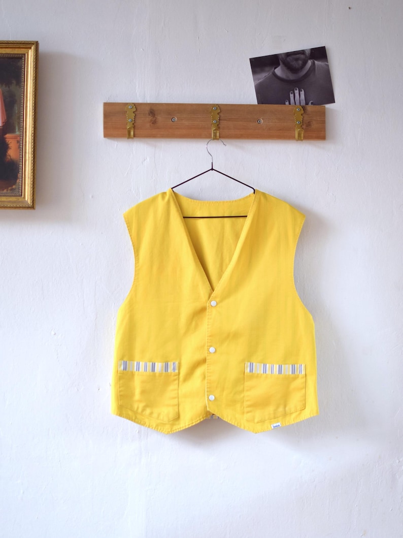May include: A bright yellow vest with a V-neck and three white buttons. The vest has two front pockets with blue and white striped accents. The vest is hanging on a black hanger against a white wall.