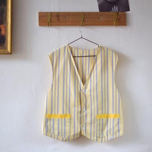 May include: A yellow and blue striped vest with two yellow pockets. The vest is hanging on a hanger against a white wall. The vest has a V-neck and button closure.