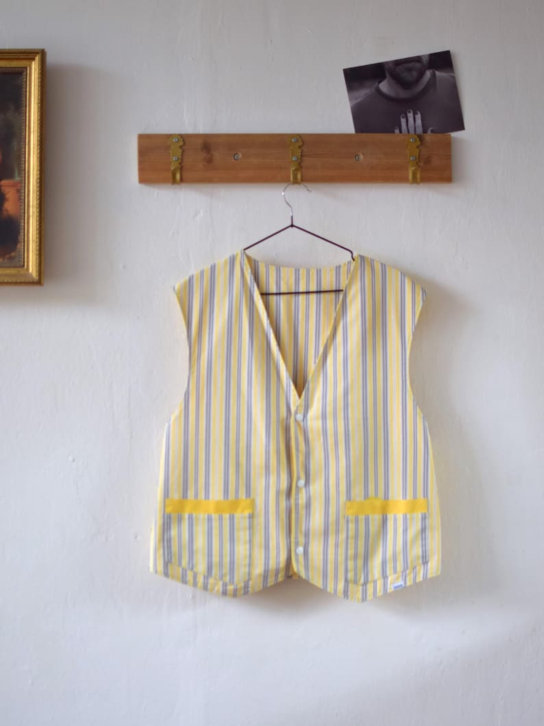 May include: A yellow and blue striped vest with two yellow pockets. The vest is hanging on a hanger against a white wall. The vest has a V-neck and button closure.