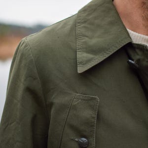 May include: A close-up of a green jacket with a buttoned collar and a flap pocket. The jacket has a casual, utilitarian style.
