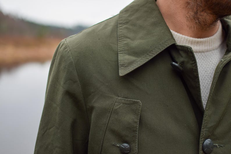 May include: A close-up of a green jacket with a buttoned collar and a flap pocket. The jacket has a casual, utilitarian style.