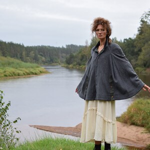 May include: A woman wearing a gray hooded cape with a brown trim and a white skirt stands in front of a river and trees.