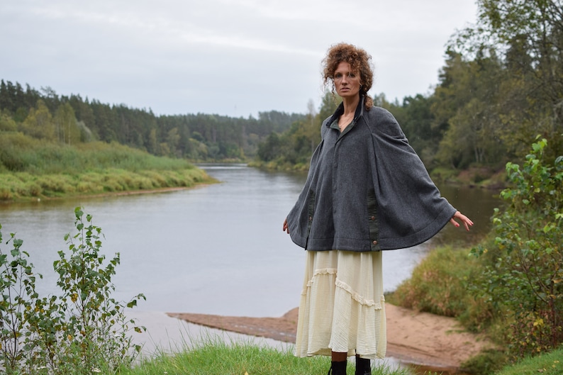 May include: A woman wearing a gray hooded cape with a brown trim and a white skirt stands in front of a river and trees.