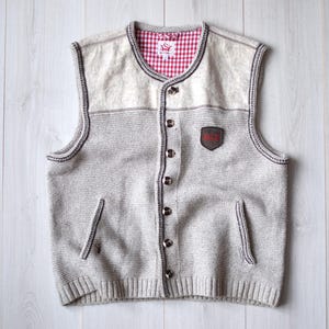 Könnte beinhalten: Eine graue Strickweste mit Knopfleiste und rotem und weißem Karomuster. Die Weste hat zwei Taschen und einen kleinen Lederfleck auf der Vorderseite.