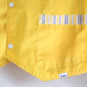 May include: Close-up of a bright yellow vest with two white buttons and two pockets. The pockets are trimmed with a striped pattern in blue, white, and yellow. The vest has a small label with the word "boca".