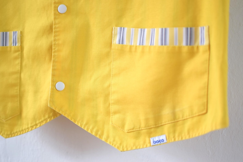 May include: Close-up of a bright yellow vest with two white buttons and two pockets. The pockets are trimmed with a striped pattern in blue, white, and yellow. The vest has a small label with the word "boca".
